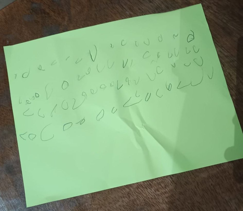 A sheet of green paper filled with rows of small invented symbols drawn in pencil. None of them are real letters.